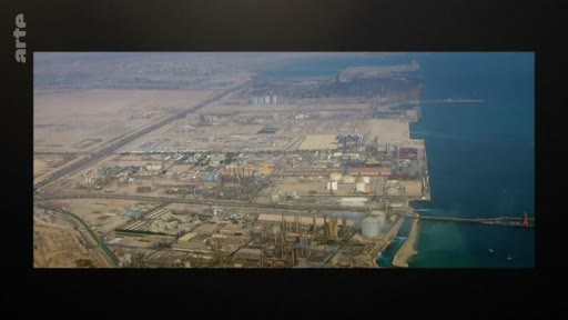 Vue aérienne panoramique d'un complexe industriel et portuaire situé en bordure de mer, à proximité d'une vaste zone désertique. L'image montre les infrastructures de production et les installations de stockage d'énergie dans un environnement aride.