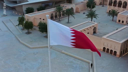 Vue aérienne du drapeau national du Qatar flottant au vent dans une cour intérieure de Doha, entourée d'une architecture traditionnelle et moderne.