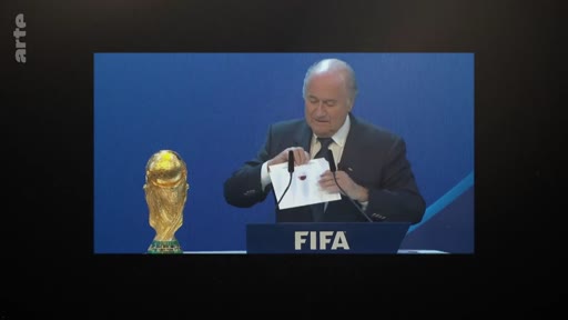 Sepp Blatter, ancien président de la FIFA, est filmé en train d'ouvrir une enveloppe lors d'une cérémonie officielle de la FIFA, avec le trophée de la Coupe du Monde visible à ses côtés.