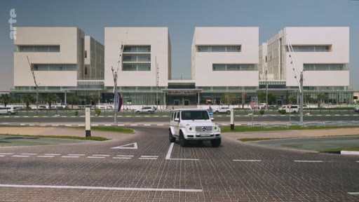 Un 4x4 Mercedes-Benz roule sur une route pavée devant un grand complexe de bâtiments modernes à l'architecture géométrique au Qatar, illustrant le développement urbain du pays.