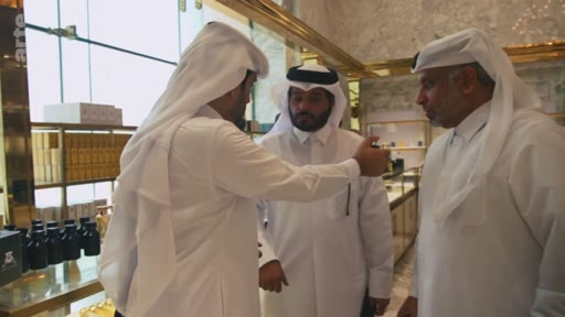Trois hommes portant le thobe et la ghutra traditionnelle qatarienne examinent des produits dans une boutique de luxe, probablement une parfumerie, au Qatar. La scène montre une interaction commerciale dans un cadre haut de gamme.