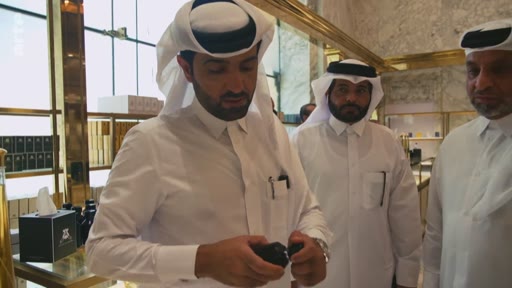 Trois hommes portant la thobe et le ghutra examinent un flacon de parfum ou un produit de luxe dans une boutique haut de gamme au Qatar. La scène illustre le commerce de luxe et les traditions vestimentaires locales.