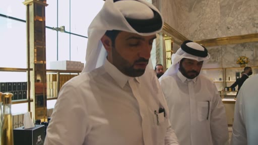 Des hommes portant la thobe et le ghutra visitent une boutique de luxe, probablement une parfumerie, dans un environnement élégant avec des décorations dorées. La scène montre des clients dans un espace commercial haut de gamme au Qatar.