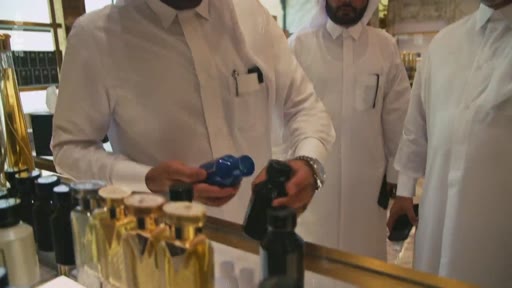 Des hommes portant des thobes traditionnelles examinent des flacons de parfum dans une boutique de luxe. La scène montre une interaction commerciale centrée sur la parfumerie, probablement au Moyen-Orient.