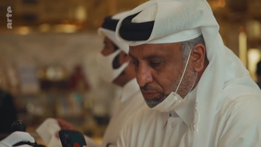 Un homme d'âge mûr portant une thobe blanche et une ghutra avec un agal noir, équipé d'un masque chirurgical, est filmé en gros plan dans un environnement intérieur. L'image illustre le port du masque et les traditions vestimentaires au Qatar.