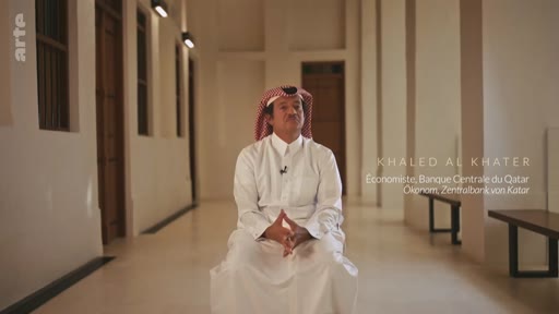 Khaled Al Khater, économiste à la Banque centrale du Qatar, est filmé en plan moyen lors d'une interview dans un couloir à l'architecture traditionnelle. Il porte une tenue traditionnelle qatarienne.