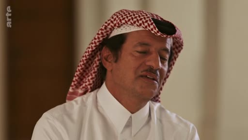 Un homme d'origine arabe, vêtu d'une chemise blanche et d'une ghutra à motifs rouges et blancs, s'exprime lors d'une interview télévisée.