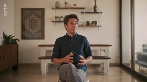 Un homme s'exprime lors d'une interview dans un intérieur domestique moderne. Il est assis sur une chaise, les mains jointes, devant une table en bois et des étagères décoratives.