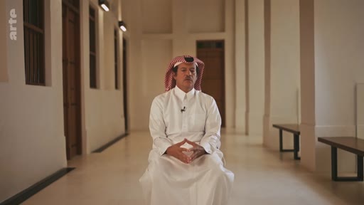 Un homme portant une thobe blanche et un keffieh rouge et blanc est assis dans un couloir aux murs blancs, s'exprimant lors d'une interview télévisée. Le décor minimaliste suggère un cadre institutionnel ou culturel au Moyen-Orient.