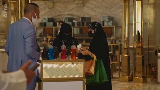 Des clients, dont des femmes portant des abayas, font des achats dans une boutique de parfums de luxe, probablement aux Émirats arabes unis. Un homme en costume bleu et masque chirurgical est également présent dans le magasin.