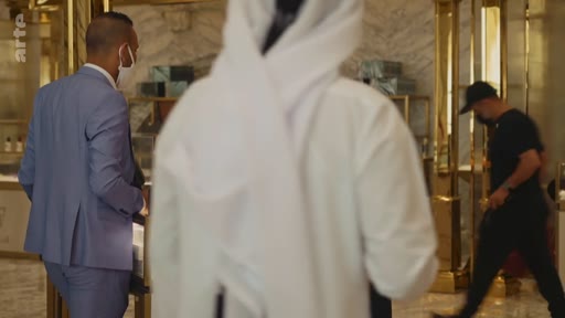 Des clients, dont un homme en tenue traditionnelle, et du personnel portant des masques chirurgicaux dans une boutique de luxe aux finitions dorées.
