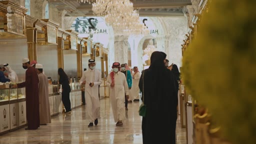 Des clients, dont certains portent des tenues traditionnelles comme le thawb et la ghutra, marchent dans les allées d'un centre commercial luxueux avec des lustres et des décorations dorées. Le lieu semble être un souk moderne ou un centre commercial haut de gamme au Moyen-Orient.