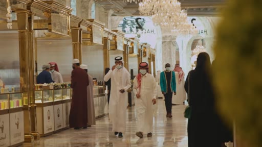 Des hommes en thobe et des femmes en abaya circulent dans les allées d'un centre commercial haut de gamme, caractérisé par une architecture dorée et des lustres imposants. L'ambiance évoque le commerce de luxe dans un pays du Golfe.