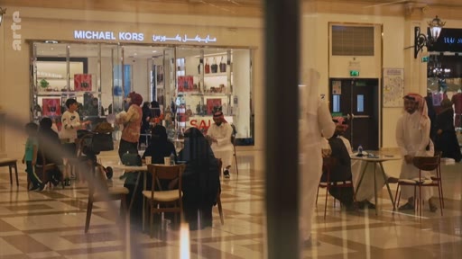 Des clients, dont certains portent des vêtements traditionnels, sont attablés dans un centre commercial devant la boutique Michael Kors. La scène illustre la vie quotidienne et la consommation dans un centre commercial au Moyen-Orient.
