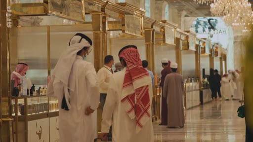 Des hommes portant des thobes et des keffiehs marchent dans les allées d'un centre commercial haut de gamme, probablement aux Émirats arabes unis ou en Arabie saoudite, caractérisé par des décorations dorées et des lustres imposants.