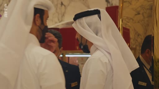 Deux hommes portant la thobe et la ghutra discutent dans un lieu public, probablement un centre commercial ou un espace de luxe, au Qatar. Ils portent des masques de protection faciale, suggérant une période liée à la pandémie de COVID-19.