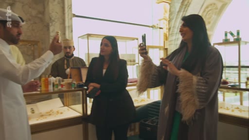 Un homme en tenue traditionnelle présente des produits de parfumerie à deux femmes dans une boutique élégante, tandis que l'une d'elles filme la scène avec son smartphone. La scène se déroule dans un espace de vente haut de gamme, probablement au Moyen-Orient.