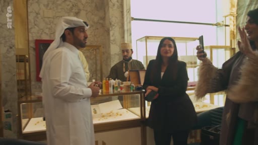 Un homme portant une thobe et un ghutra discute avec une femme en blazer noir et d'autres personnes dans une boutique de luxe, probablement une bijouterie ou une parfumerie, à Doha au Qatar. Une personne filme la scène avec un smartphone.