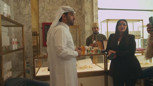 Un homme portant une thobe et un ghutra discute avec une femme en blazer noir dans une boutique de luxe, probablement une parfumerie ou une bijouterie, lors d'un reportage télévisé. En arrière-plan, un autre homme en tenue traditionnelle travaille derrière un comptoir vitré.