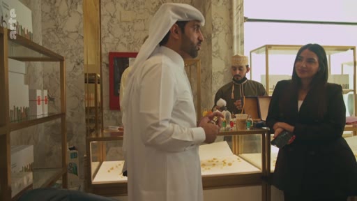 Un homme portant une thobe et un ghutra discute avec une femme en tenue professionnelle dans une boutique de luxe, probablement à Doha, au Qatar. La scène montre une interaction commerciale ou une interview dans un environnement haut de gamme avec des vitrines de bijoux ou de parfums.
