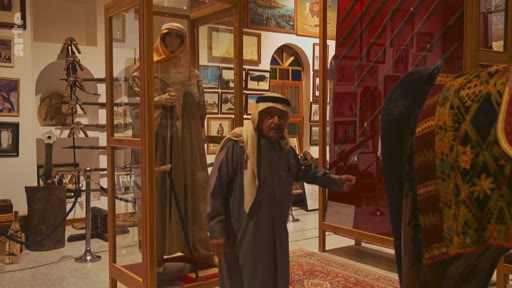 Un homme âgé portant une ghutra et une thobe visite un musée local, probablement dans la péninsule arabique, où sont exposés des artefacts historiques, des armes anciennes et des mannequins en costume traditionnel. Le cadre met en valeur le patrimoine culturel et l'histoire régionale à travers des vitrines d'exposition.