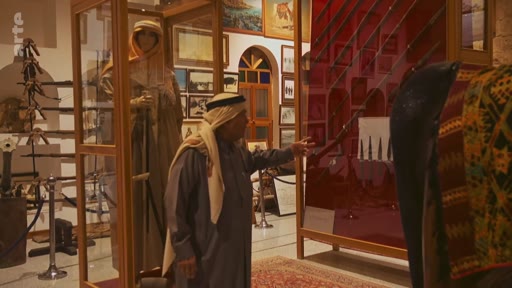 Un homme portant une thobe et un keffieh visite un musée local, observant des vitrines contenant des artefacts historiques, des armes anciennes et des photographies d'époque. La scène illustre la préservation du patrimoine culturel et historique dans une région du Moyen-Orient.