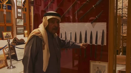 Un homme âgé portant une ghutra et un thobe présente une vitrine contenant des poignards traditionnels dans un musée ou une exposition historique. La scène illustre la préservation du patrimoine culturel et des objets artisanaux dans une région du Moyen-Orient.