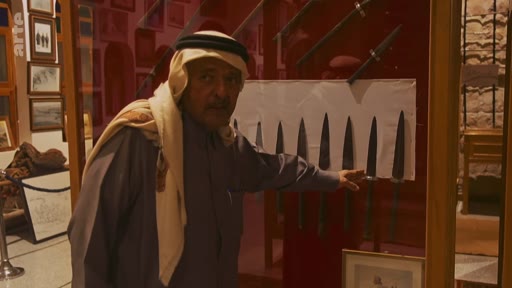 Un homme portant une tenue traditionnelle et un keffieh présente une collection de poignards exposés derrière une vitrine dans un musée. Il pointe du doigt l'un des objets historiques lors d'une visite guidée.