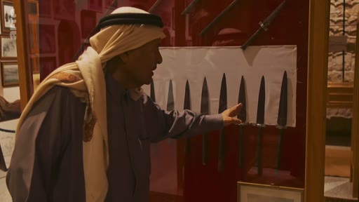 Un homme portant une ghutra et un thobe examine une vitrine contenant des poignards traditionnels dans un musée en Arabie saoudite. Il pointe du doigt l'un des objets exposés lors de cette visite culturelle.