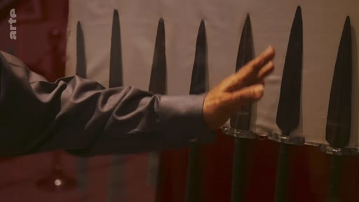 Une main humaine passe devant une rangée de pointes de lances métalliques exposées, probablement dans un musée ou une collection d'armes historiques.