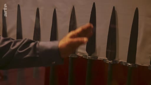 Une main humaine pointe vers une collection de pointes de lances en métal exposées sur un support, illustrant une présentation historique ou archéologique.