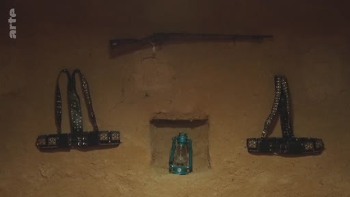 Vue d'intérieur montrant un fusil, deux cartouchières et une lampe à pétrole disposés sur un mur en terre battue, évoquant une esthétique traditionnelle ou historique.