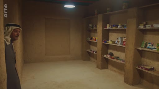 Un homme portant une ghutra et un thobe visite une pièce rustique aux murs en terre battue, équipée d'étagères de rangement contenant divers produits, dans un contexte de reportage sur le patrimoine ou la vie traditionnelle.