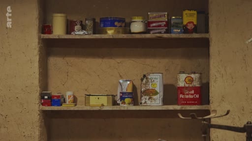 Des étagères en bois rustiques dans une pièce aux murs en terre battue, exposant divers produits de consommation, notamment des boîtes de conserve, des bidons d'huile Shell Rotella Oil et des emballages vintage.