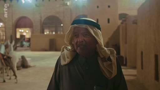 Un homme âgé vêtu d'une thobe et d'un ghutra traditionnel se tient dans un espace extérieur aux murs en terre cuite, évoquant l'architecture traditionnelle de l'Arabie saoudite.
