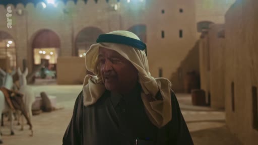 Un homme âgé vêtu d'une thobe et d'un keffieh avec un agal, se tient devant une architecture traditionnelle en terre cuite dans un village historique, probablement dans la péninsule arabique.