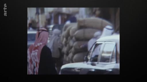 Des habitants en tenue traditionnelle circulent dans une rue du Qatar au début des années 1970, à l'époque de l'indépendance du pays. On aperçoit des hommes en ghutra et des sacs empilés près d'une voiture dans un décor urbain typique de cette période.