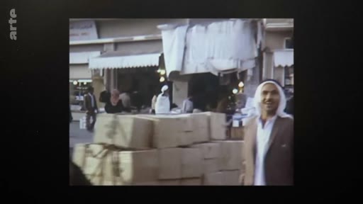Un homme portant une ghutra et un thobe sourit devant une boutique dans une rue commerçante au Qatar, lors d'images d'archives illustrant l'indépendance du pays en 1971. Des cartons sont empilés au premier plan, témoignant de l'activité commerciale de l'époque.