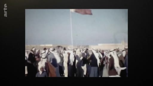 Des hommes en tenue traditionnelle qatarienne sont rassemblés en extérieur lors d'une cérémonie officielle, avec un drapeau flottant au-dessus de la foule. Cette image d'archive illustre le contexte politique et social du Qatar avant la révolution de palais de 1995.
