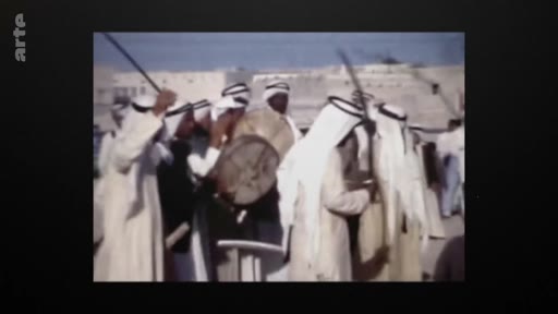 Des hommes en tenue traditionnelle arabe, portant des keffiehs, participent à une danse folklorique traditionnelle avec des bâtons et un tambour lors d'une célébration publique.