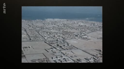 Vue aérienne panoramique d'une ville côtière située dans un environnement aride ou désertique, probablement au Moyen-Orient ou en Afrique du Nord.