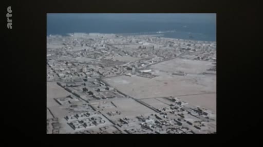 Vue aérienne panoramique d'une ville située en bord de mer, entourée par un paysage aride et désertique. L'image montre l'urbanisation dense le long du littoral.