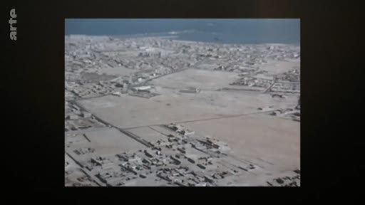 Vue aérienne d'une ville côtière avec des zones désertiques et des quartiers résidentiels, probablement dans une région aride du Moyen-Orient ou d'Afrique du Nord.