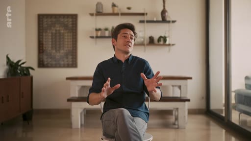 Un homme s'exprime lors d'une interview dans un intérieur domestique, les mains en mouvement pour appuyer ses propos.