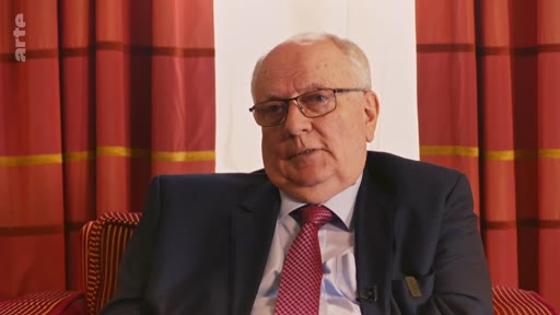 Un homme âgé, portant des lunettes, un costume bleu et une cravate rouge, est interviewé dans un cadre formel avec des rideaux en arrière-plan. Il s'agit probablement d'une personnalité politique ou d'un expert s'exprimant sur un sujet d'actualité.