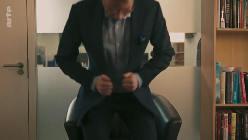 Un homme en costume et chemise ajuste sa veste tout en étant assis sur un fauteuil dans un bureau, avec une bibliothèque visible en arrière-plan.