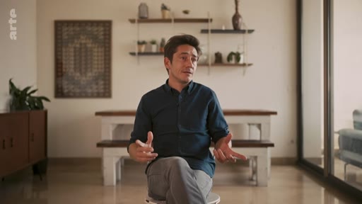 Un homme s'exprime face caméra lors d'une interview dans un intérieur moderne. Il porte une chemise bleu foncé et est assis sur un tabouret, les mains en mouvement pour appuyer ses propos.