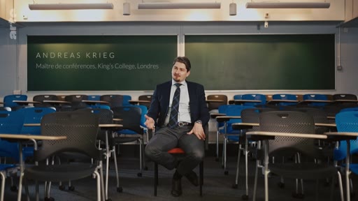Andreas Krieg, maître de conférences au King's College de Londres, est assis dans une salle de classe vide lors d'une interview télévisée. Il porte un costume et une cravate, et le texte à l'écran identifie son nom et sa fonction académique.
