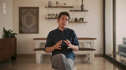 Un homme s'exprime lors d'une interview dans un salon à l'aménagement contemporain, avec des étagères murales et une table en arrière-plan.