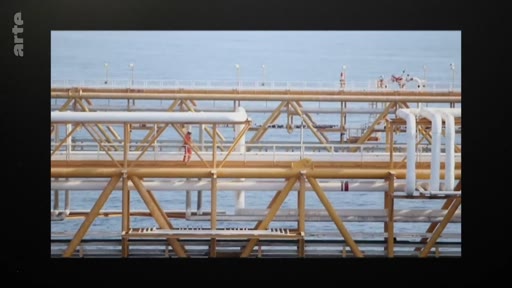 Un ouvrier en tenue de travail rouge marche sur une passerelle métallique au-dessus de la mer, sur une plateforme pétrolière ou gazière offshore. Cette image illustre les activités industrielles en milieu marin et l'exploitation des ressources énergétiques.
